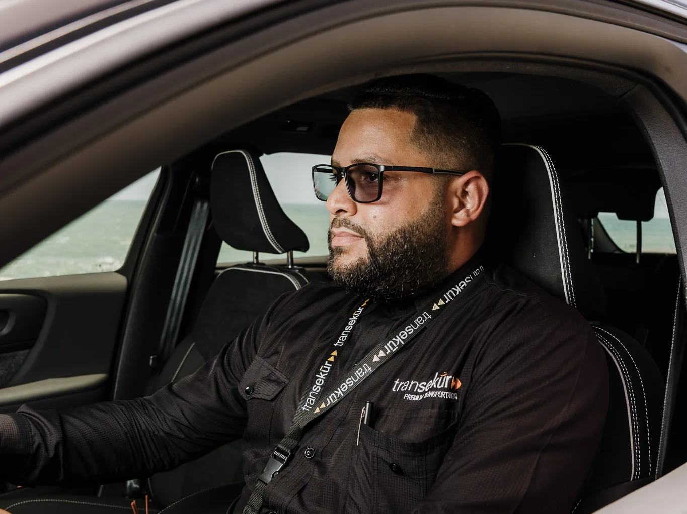 A Transekur chauffeur at the wheel of a white Volvo XC40 Recharge, with the sea behind