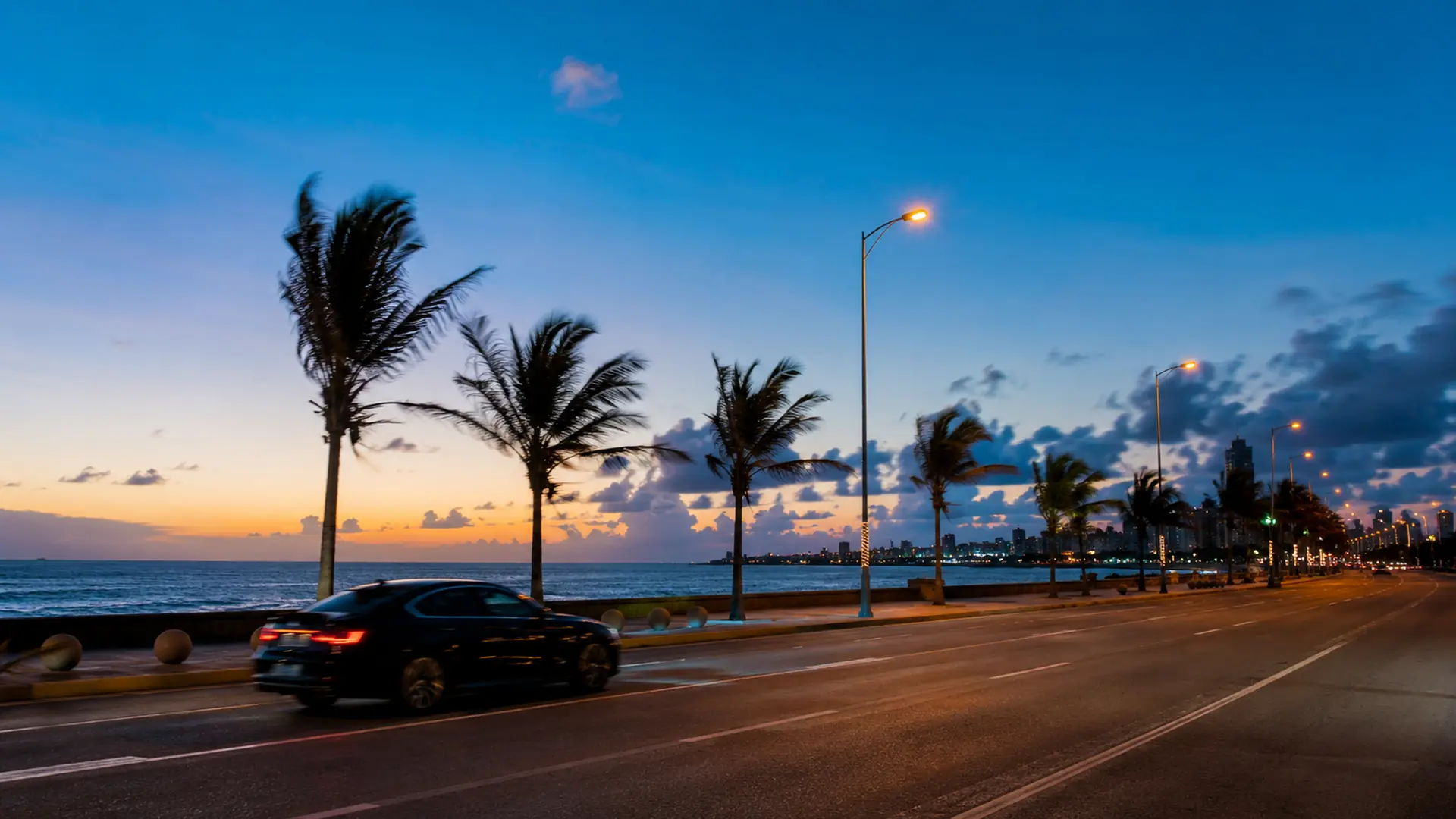 A car passing wind-bent palms on the Malecón seafront road in Santo Domingo at nightfall