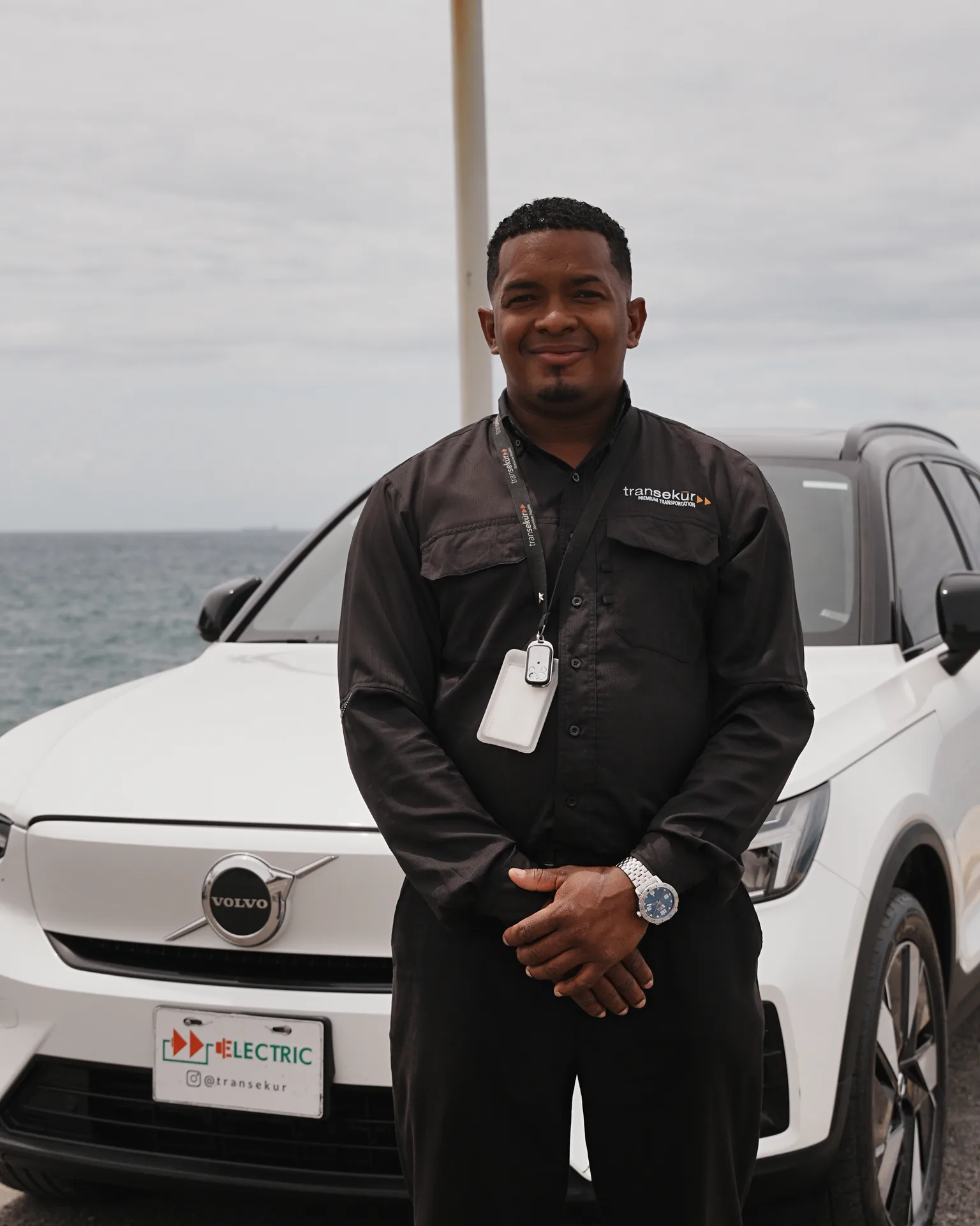 A uniformed Transekur chauffeur standing in front of a white Volvo XC40 Recharge with Transekur ELECTRIC plates, the sea behind