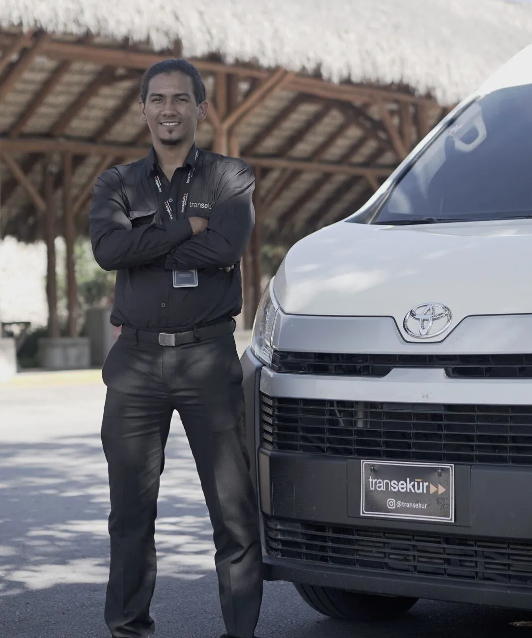 A Transekur chauffeur in a black uniform with arms crossed beside a white Toyota van with Transekur plates