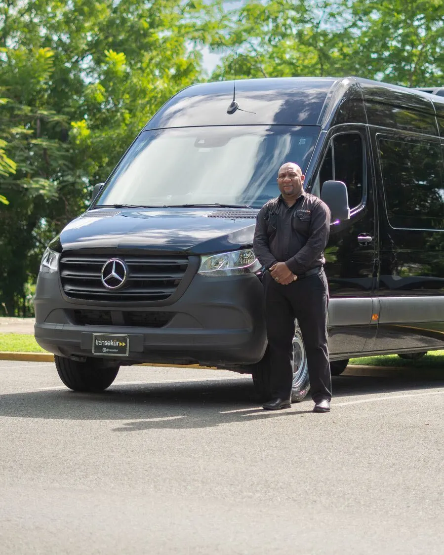 A uniformed Transekur chauffeur standing beside a black Mercedes Sprinter van with Transekur plates, green trees behind