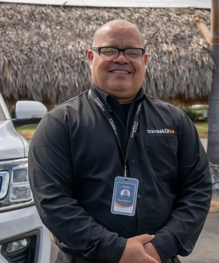 A smiling Transekur chauffeur in a black uniform with company ID badge beside a white SUV under a thatched canopy
